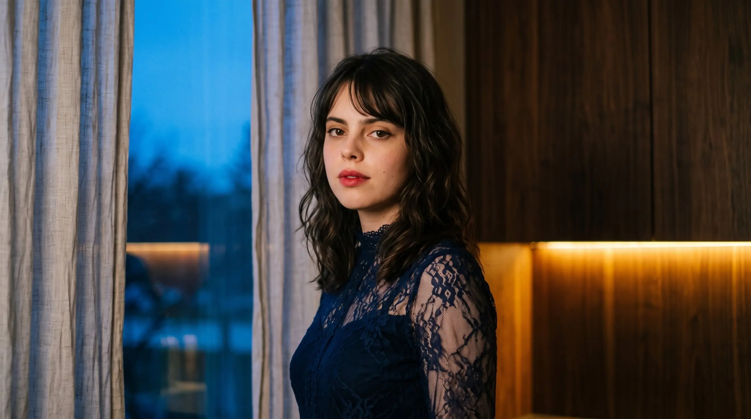 Lily in navy lace by a window at blue hour, lit by warm lamp light
