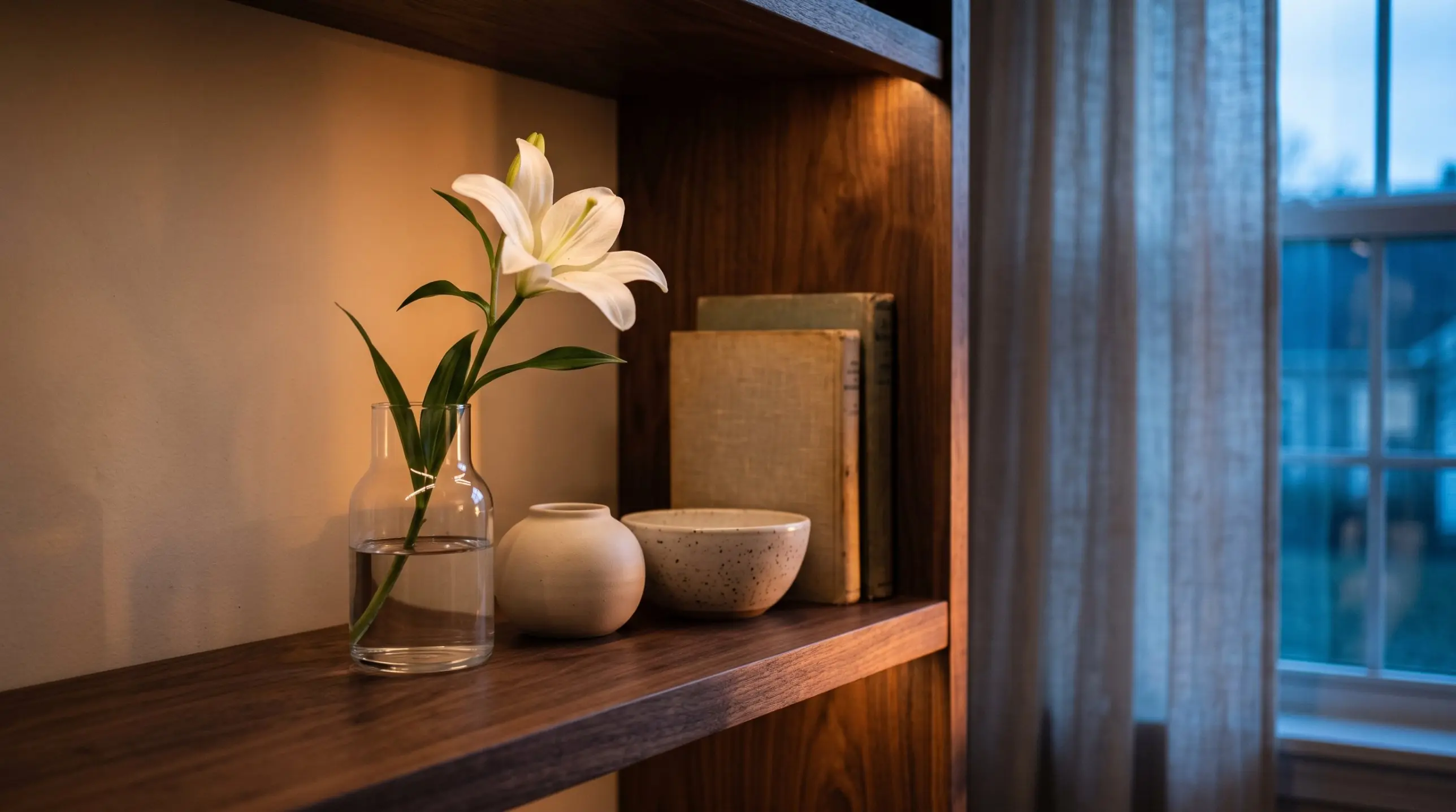 A single white lily in a glass vase on a walnut shelf, lit by warm lamp light at blue hour